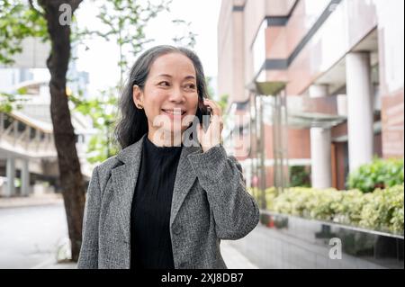 Eine schöne, positive, reife asiatische Geschäftsfrau telefoniert am Telefon, während sie ihre Arbeit pendelt und auf einem Bürgersteig in der Stadt läuft. Geschäftsleute Stockfoto