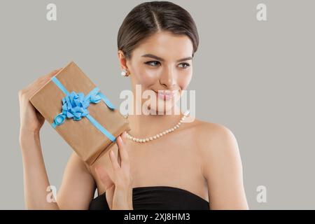Glückliche lächelnde Frau, die beige Geschenkbox über grauem Hintergrund hält, Nahaufnahme Porträt des Schönheitsmodestudios Stockfoto