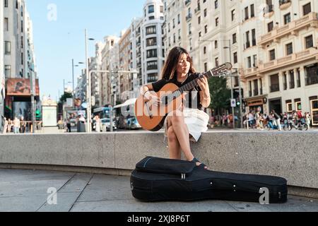 Junge Musikstudentin sitzt in der Stadt und spielt Akustikgitarre. Vollständige Ansicht einer lateinischen Frau mit einer Gitarre in der Hand und dem Gehäuse auf dem Boden. Stockfoto