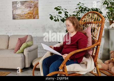 Ein kleines Mädchen mit Down-Syndrom sitzt in einem Schaukelstuhl und liest nachdenklich ein Buch. Stockfoto