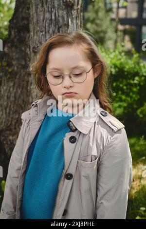 Ein kleines Mädchen mit Down-Syndrom steht an einem Baum, verloren in Gedanken. Stockfoto