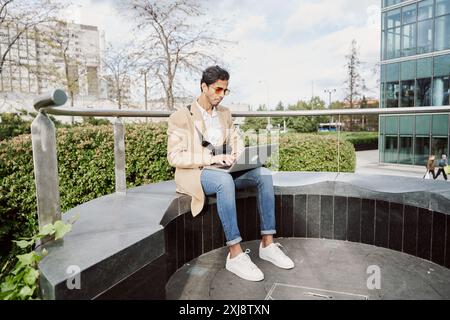 Vorderansicht eines jungen Mannes aus dem Nahen Osten, der im Freien mit Laptop arbeitet, online arbeitet oder im Internet surft, Kopierraum. Ein Indianer sitzt auf der Bank auf der Straße und tippt Stockfoto