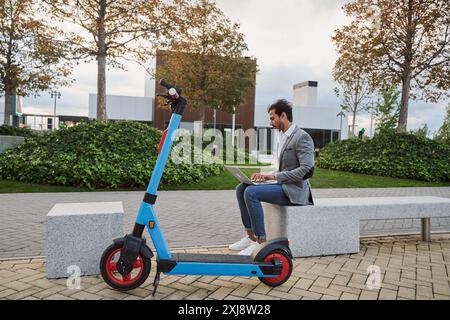 Junger Geschäftsmann, der seinen Laptop benutzt und auf einer Steinbank sitzt, mit dem Elektroroller neben ihm und Bürogebäuden im Hintergrund Stockfoto