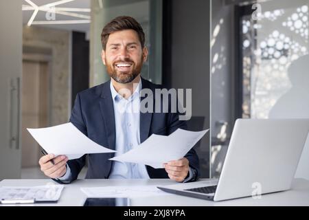 Porträt eines glücklichen jungen Büroarbeiters, eines Geschäftsmannes, der in einem Anzug am Schreibtisch sitzt, lächelt und Dokumente und eine Zustimmung zur Kamera zeigt. Stockfoto