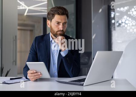 Seriöser, bärtiger Geschäftsmann in einem Anzug, der am Schreibtisch sitzt, während er einen Laptop benutzt und ein Tablet in einer hellen, modernen Büroumgebung hält. Stockfoto