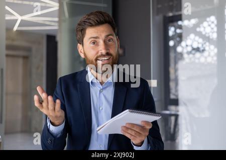 Dieses Foto zeigt einen selbstbewussten, professionellen Mann, der Ideen in einer modernen Büroumgebung leidenschaftlich diskutiert oder präsentiert. Er hält Notizen und spricht mit seinem Publikum. Stockfoto