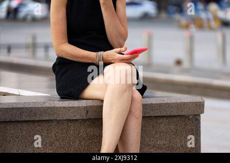 Frau mit überkreuzten Beinen, die ein kurzes Sommerkleid trägt und mit Smartphone auf der Straßenbank von а sitzt Stockfoto
