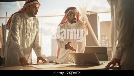 Erfolgreiche muslimische Geschäftsleute in traditionellen weißen Outfits, die ein Büro treffen, verhandeln und über finanzielle Chancen sprechen. Laptop Verwenden. Saudi, Emirat, Arabisches Geschäftsmann-Konzept. Stockfoto