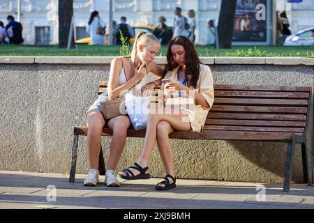 Zwei hübsche Mädchen sitzen mit Smartphones auf einer Straße. Mit dem Mobiltelefon in der Sommerstadt Stockfoto