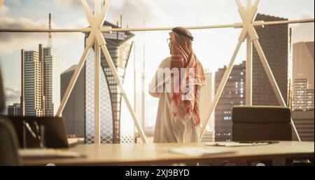Erfolgreicher muslimischer Geschäftsmann in traditionellem weißem Outfit in seinem modernen Büro mit Blick aus dem Fenster auf Big City mit Wolkenkratzern. Erfolgreiches Konzept Für Saudi-Arabien, Emirate, Arabische Geschäftsleute Stockfoto