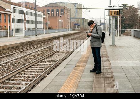 Ein Mann überprüft sein Handy, während er auf einem Bahnsteig steht und auf einen Zug wartet. Stockfoto