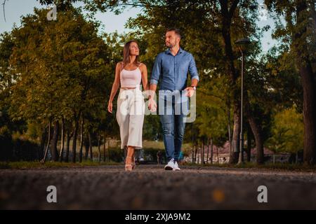 Ein junges Paar geht Hand in Hand durch einen von Bäumen gesäumten Park. Die Sonne untergeht im Hintergrund und strahlt ein warmes Licht auf die Szene. Stockfoto