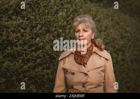 Eine ältere Frau steht in einem Garten mit grünem Laub, trägt einen braunen Trenchcoat und einen orangefarbenen Schal. Stockfoto