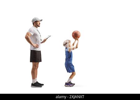 Profilaufnahme eines Trainers in voller Länge, der einen Jungen beobachtet, der einen Basketball auf weißem Hintergrund schießt Stockfoto