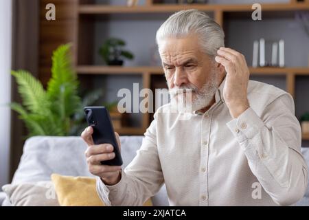 Älterer Mann mit grauen Haaren und Bart, der mit einem verwirrten Ausdruck auf sein Smartphone schaut, während er zu Hause auf einer bequemen Couch sitzt. Stockfoto