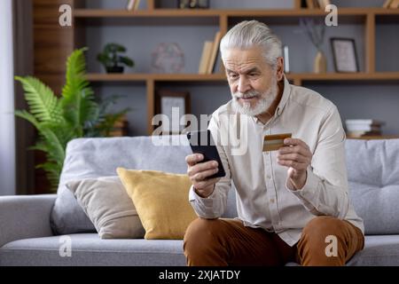 Grauhaariger verängstigter Mann verwendet Kreditkarte über mobile App, hält Telefon, sitzt zu Hause auf dem Sofa. Stockfoto