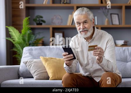 Porträt eines lächelnden älteren Mannes zu Hause, der auf der Couch sitzt und in die Kamera blickt. Enthält eine Kreditkarte und ein Mobiltelefon. Stockfoto