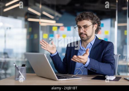 Mann mit Anzug Brille engagierte Videokonferenzanruf mit Laptop und Kopfhörern in moderner Büroumgebung, besprich Arbeit mit ausdrucksstarker Hand g Stockfoto