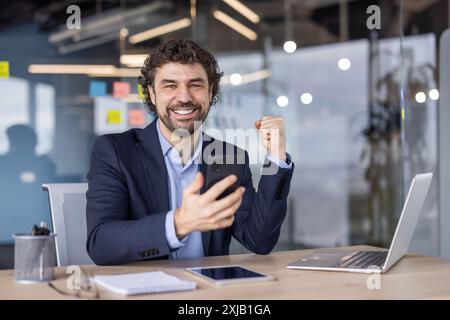 Fröhlicher Geschäftsmann, der Erfolg im modernen Büro feiert. Mann im Anzug, der Smartphone hält, lächelt, eine Faustpumpe macht. Unternehmenskonzept achi Stockfoto