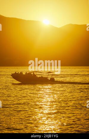 Ein Motorboot mit Leuten, die während des Sonnenuntergangs entlang der Bucht segeln. Goldenes Meer und Berge an einem Sommerabend in der Bucht von Kotor, Montenegro Stockfoto