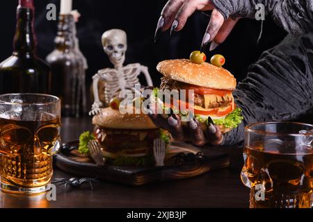 Die Hexenhände halten einen Monster Burger in ihren Händen auf einem sitzenden Skelett. Die perfekte Vorspeise für die Halloween Party Stockfoto