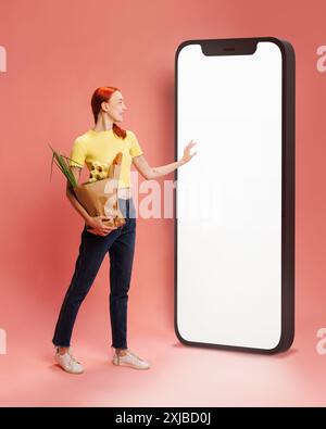 Frau in gelbem Oberteil und Jeans, die Lebensmittel hält und in ein großes Telefon mit leerem weißen Bildschirm vor rosa Hintergrund greift Stockfoto
