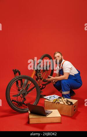 Mechaniker, bärtiger Mann in blauem Overall, der Fahrrad fixiert, umgeben von Werkzeugen und Laptop, vor rotem Hintergrund Stockfoto