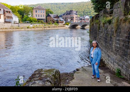 Erwachsene Frau, die entspannt am Ufer des Flusses Ourthe steht, Gebäude, Brücke und Berg mit grünen Bäumen im unscharfen Hintergrund, blaue Freizeitkleidung, sunn Stockfoto