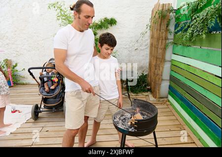 Vater und Sohn genießen einen Grillabend im Garten, während Familienmitglieder, darunter ein Baby im Kinderwagen, gute Zeit miteinander verbringen. Sommer Familienbindung o Stockfoto