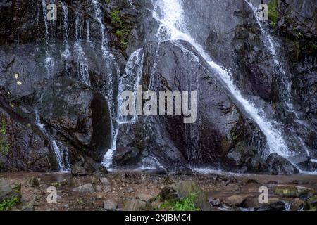 Ein lächelnder Mann steht in der Nähe eines kaskadierenden Wasserfalls auf nassen, dunklen Felsen, trägt einen Rucksack und Outdoor-Ausrüstung und genießt eine Naturwanderung Stockfoto