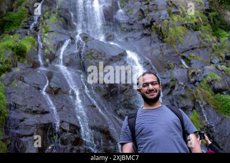 Ein lächelnder Mann steht in der Nähe eines kaskadierenden Wasserfalls auf nassen, dunklen Felsen, trägt einen Rucksack und Outdoor-Ausrüstung und genießt eine Naturwanderung Stockfoto