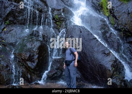 Ein lächelnder Mann steht in der Nähe eines kaskadierenden Wasserfalls auf nassen, dunklen Felsen, trägt einen Rucksack und Outdoor-Ausrüstung und genießt eine Naturwanderung Stockfoto