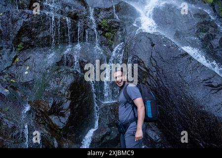 Ein lächelnder Mann steht in der Nähe eines kaskadierenden Wasserfalls auf nassen, dunklen Felsen, trägt einen Rucksack und Outdoor-Ausrüstung und genießt eine Naturwanderung Stockfoto