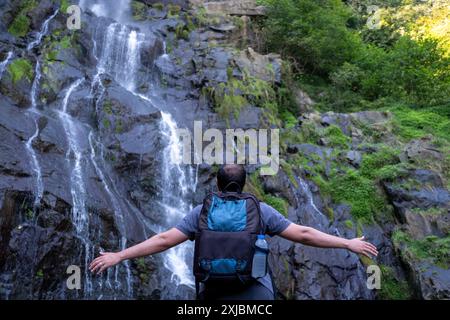 Ein lächelnder Mann steht in der Nähe eines kaskadierenden Wasserfalls auf nassen, dunklen Felsen, trägt einen Rucksack und Outdoor-Ausrüstung und genießt eine Naturwanderung Stockfoto