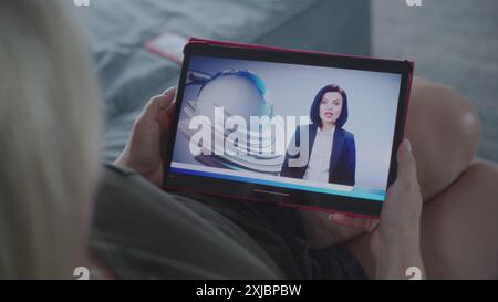 Der Tablet-Bildschirm zeigt Fernsehnachrichten mit weiblicher Nachrichtensprecherin. Kaukasische Frau, die Nachrichten über US-Präsidentschaftswahlen mit einem digitalen Tablet auf dem Sofa ansieht. Nahaufnahme. Stockfoto