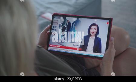 Der Tablet-Bildschirm zeigt Fernsehnachrichten mit weiblicher Nachrichtensprecherin. Kaukasische Frau, die Nachrichten über US-Präsidentschaftswahlen mit einem digitalen Tablet auf dem Sofa ansieht. Nahaufnahme. Stockfoto