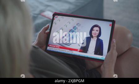 Der Tablet-Bildschirm zeigt Fernsehnachrichten mit weiblicher Nachrichtensprecherin. Kaukasische Frau, die Nachrichten über US-Präsidentschaftswahlen mit einem digitalen Tablet auf dem Sofa ansieht. Nahaufnahme. Stockfoto