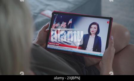 Der Tablet-Bildschirm zeigt Fernsehnachrichten mit weiblicher Nachrichtensprecherin. Kaukasische Frau, die Nachrichten über US-Präsidentschaftswahlen mit einem digitalen Tablet auf dem Sofa ansieht. Nahaufnahme. Stockfoto