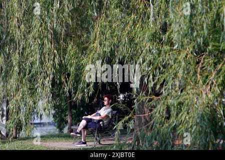 Bukarest, Rumänien. Juli 2024. Eine Frau sitzt am 17. Juli 2024 auf einer Bank im Schatten eines Parks in Bukarest, Rumänien. Die rumänische nationale Meteorologische Verwaltung (ANM) hat ihre roten und orangen Hitzewarnungen bis Mittwoch verlängert, was das gesamte Land während einer andauernden Hitzewelle betrifft. Quelle: Cristian Cristel/Xinhua/Alamy Live News Stockfoto