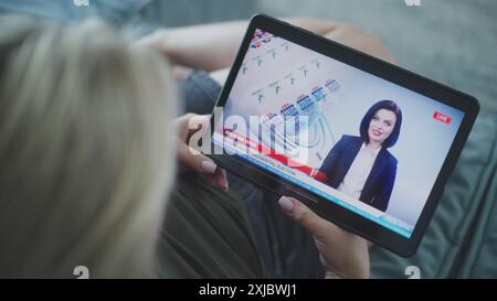 Der Tablet-Bildschirm zeigt Fernsehnachrichten mit weiblicher Nachrichtensprecherin. Kaukasische Frau, die Nachrichten über US-Präsidentschaftswahlen mit einem digitalen Tablet auf dem Sofa ansieht. Nahaufnahme. Stockfoto