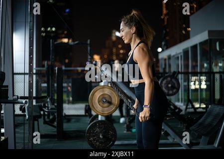 Eine Frau in schwarzer Trainingskleidung führt nachts in einem Fitnessraum im Freien eine Übung mit Kabelreihen durch. Stockfoto