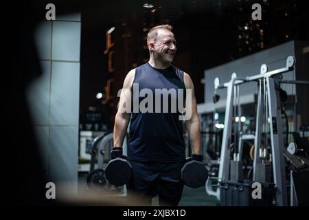 Ein Mann in einem schwarzen Tank hebt Kurzhanteln, während er nachts in einem Fitnessstudio lächelt. Stockfoto