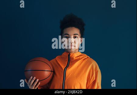 Porträt eines jungen Basketballspielers in oranger Sportkleidung, der im Studio posiert. Frau mit einem Basketball, der vor blauem Hintergrund steht. Stockfoto