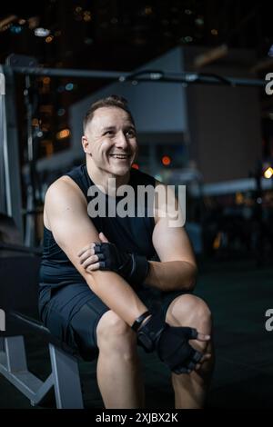 Ein Mann in schwarzer Sportkleidung sitzt auf einer Bank in einem Fitnessstudio, legt seine Arme, lächelt und trägt schwarze Handschuhe. Stockfoto