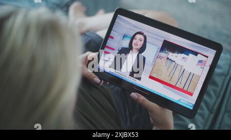 Der Tablet-Bildschirm zeigt Fernsehnachrichten mit weiblicher Nachrichtensprecherin. Kaukasische Frau, die Nachrichten über US-Präsidentschaftswahlen mit einem digitalen Tablet auf dem Sofa ansieht. Nahaufnahme. Stockfoto