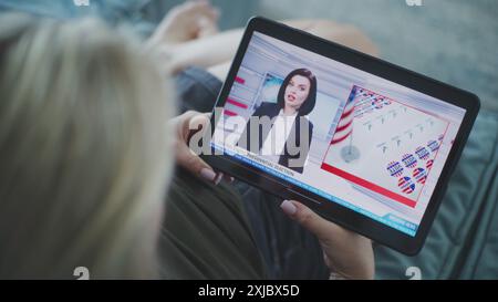 Der Tablet-Bildschirm zeigt Fernsehnachrichten mit weiblicher Nachrichtensprecherin. Kaukasische Frau, die Nachrichten über US-Präsidentschaftswahlen mit einem digitalen Tablet auf dem Sofa ansieht. Nahaufnahme. Stockfoto