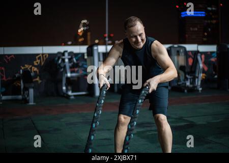 Ein Mann in einem schwarzen Tanktop und Shorts trainiert nachts mit Kampfseilen auf einem Fitnessraum auf dem Dach. Stockfoto
