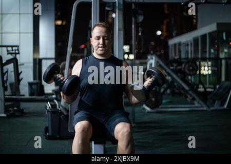 Ein Mann hebt nachts in einem Fitnessstudio Gewichte. Er trägt ein schwarzes Tanktop und Shorts. Stockfoto