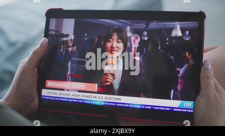 Tablet-Bildschirm zeigt die Wiedergabe von Nachrichten mit asiatischen Korrespondenten. Die kaukasische Frau sieht Nachrichtenberichte über US-Wahlen mit einem Tablet-Computer, während sie auf dem Sofa liegt. Nahaufnahme. Stockfoto