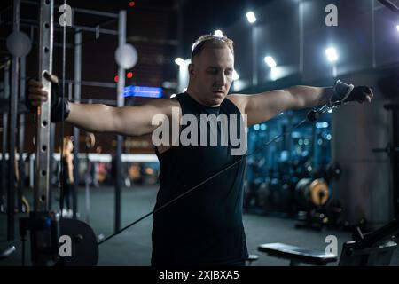 Ein Mann in einem schwarzen Tanktop trainiert nachts mit einem Kabelgerät in einem Fitnesscenter. Stockfoto